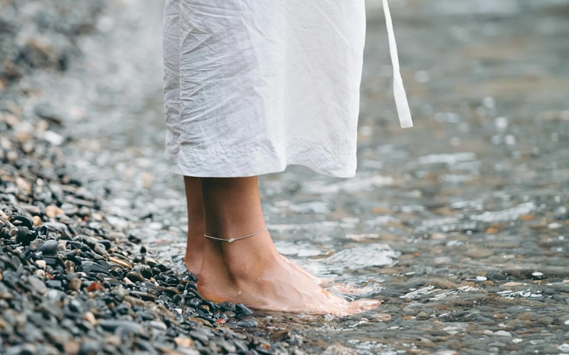 Person standing barefoot at edge of water on pebbly shore.