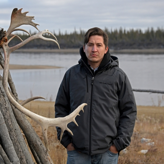 man in heavy jacket stands next to caribou antlers with water and trees in background