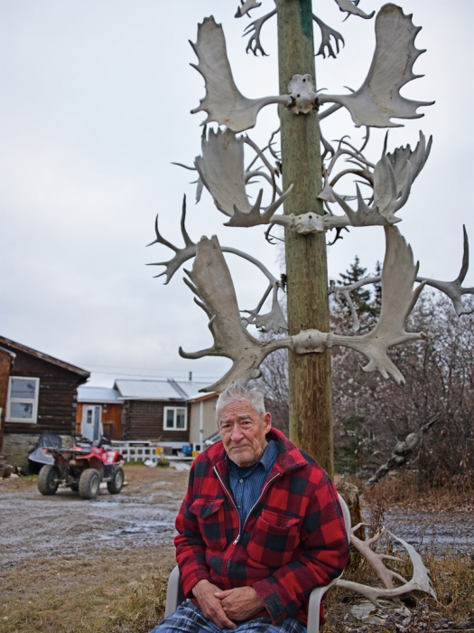 Man with gray hair and red plaid flannel jacket seated in front of tall pole with multiple caribou antlers