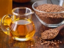 brown flaxseed in clear glass bowl and on wooden spoon and small glass pitcher of flaxseed oil
