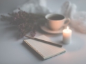 Softly lit image of an open journal with pen, cup of coffee, lit candle, and flowers on the table.