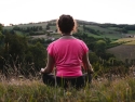 Woman in a field meditating, overlooking hills and a valley.