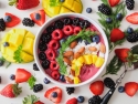 bowl of yogurt topped with blackberries, raspberries, nuts, pineapple surrounded by blackberries, blueberries, strawberries, pineapple and sprigs of parsley on table